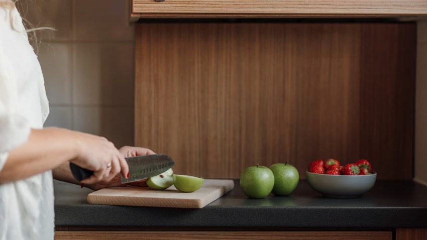 Side view of crop unrecognizable female in white shirt cutting green apples on wooden chopping board placed near bowl of sweet strawberries while preparing healthy breakfast in kitchen